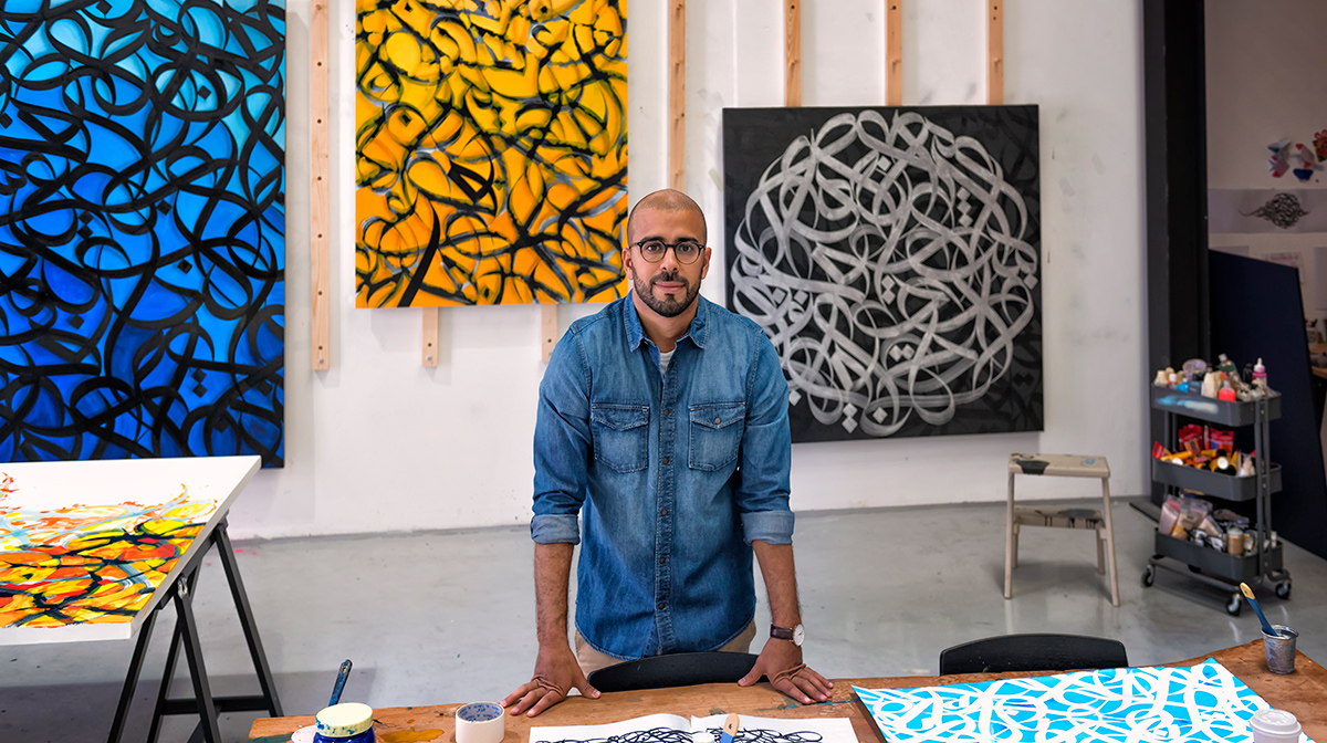 eL Seed stands in front of several of his colourful, and abstract artworks that are hung on a wall, and behind a workbench